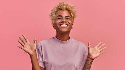 Mensch mit blonder Afro-Frisur streckt erfreut die Arme zur Seite