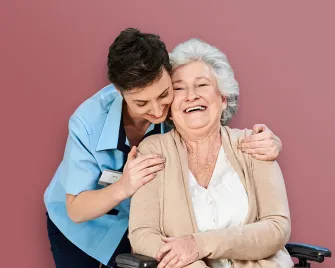 Pflegende Person umarmt eine sitzende ältere Frau